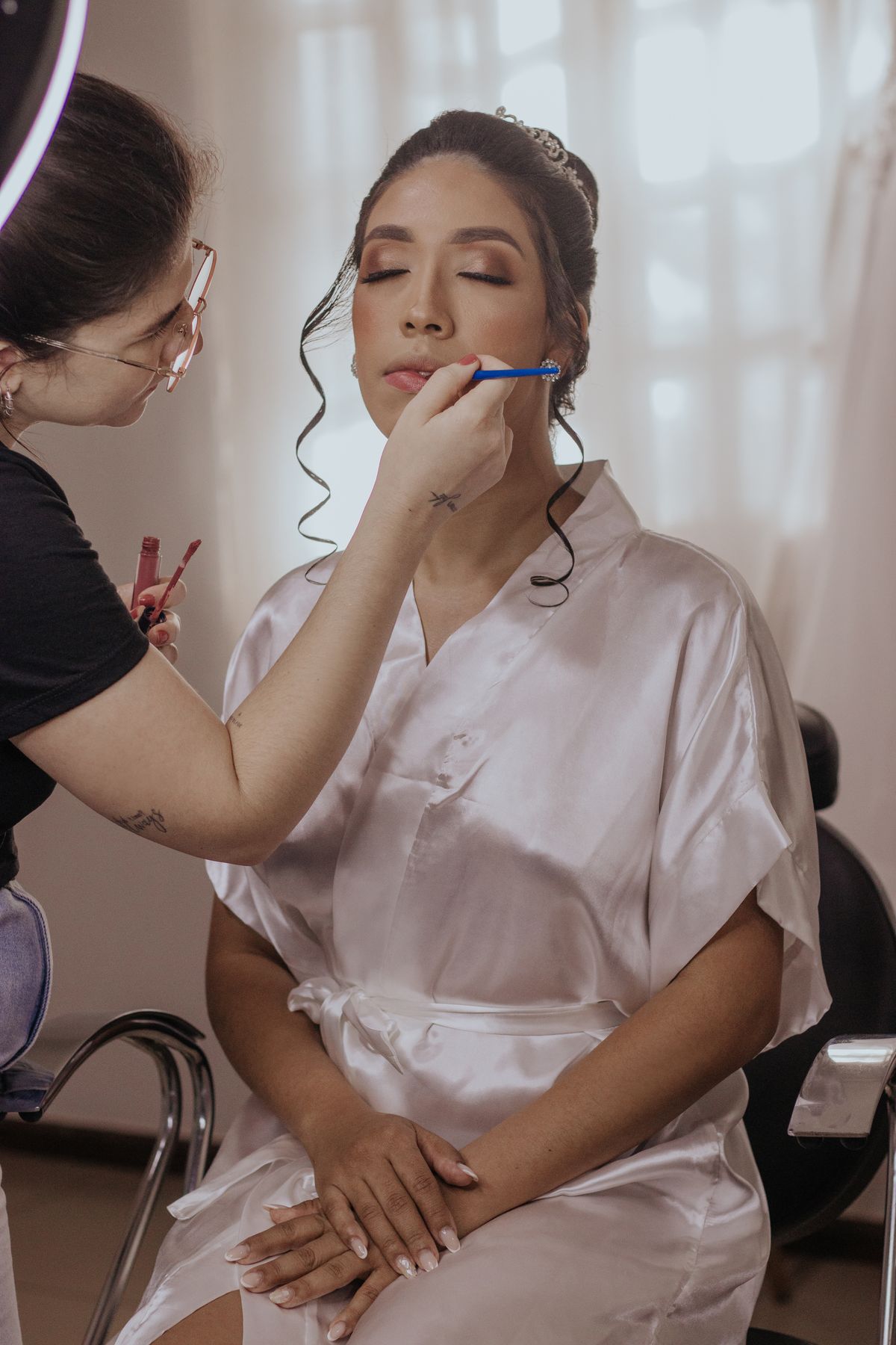 A bride sits with eyes closed, receiving elegant makeup by a beautician indoors, preparing for her wedding.