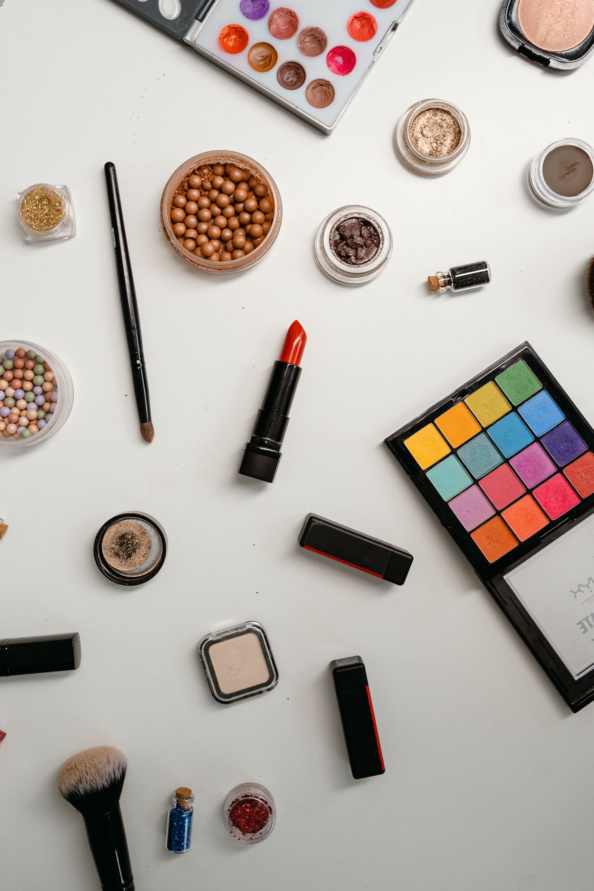 Flat lay of diverse makeup items including eyeshadow palettes, lipstick, and brushes on a white background.